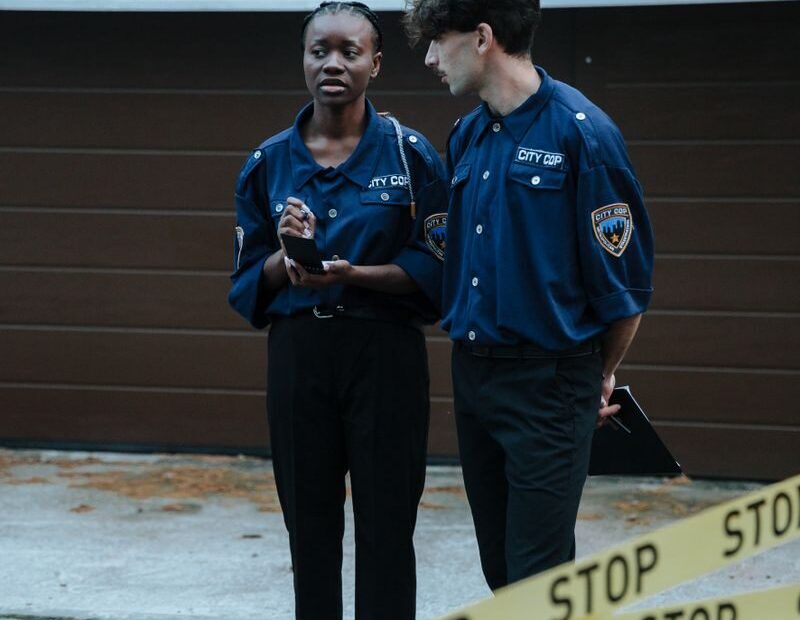 Two police officers standing behind barrier tape in front of a garage, investigating a crime scene outdoors.