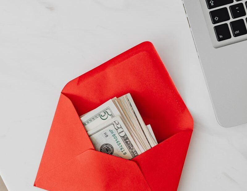 Top view of dollar bills in a red envelope next to a laptop on a white desk.