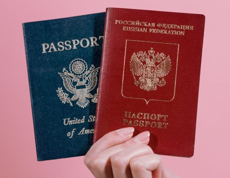 A hand holding US and Russian passports against a pink background.