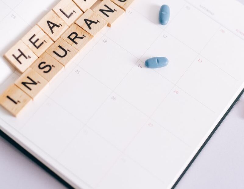 Scrabble tiles spelling 'health insurance' on a calendar beside blue pills, symbolizing medication schedule.