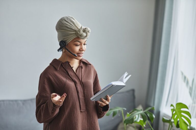 Positive African American female call agent with microphone speaking while reading notes in notepad during work in light room at home