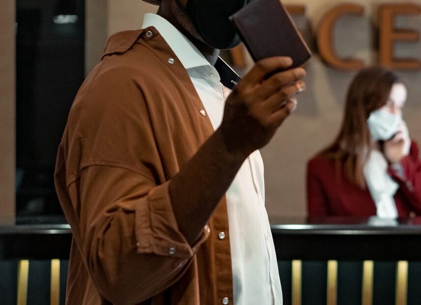 A man wearing a mask and hat holds a passport at a hotel reception, emphasizing travel safety.